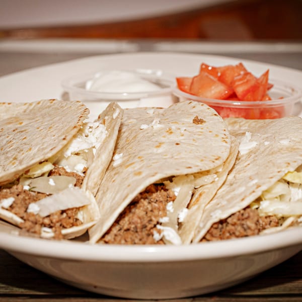 Three soft tacos filled with ground meat, cheese, and lettuce, served with sour cream and diced tomatoes.