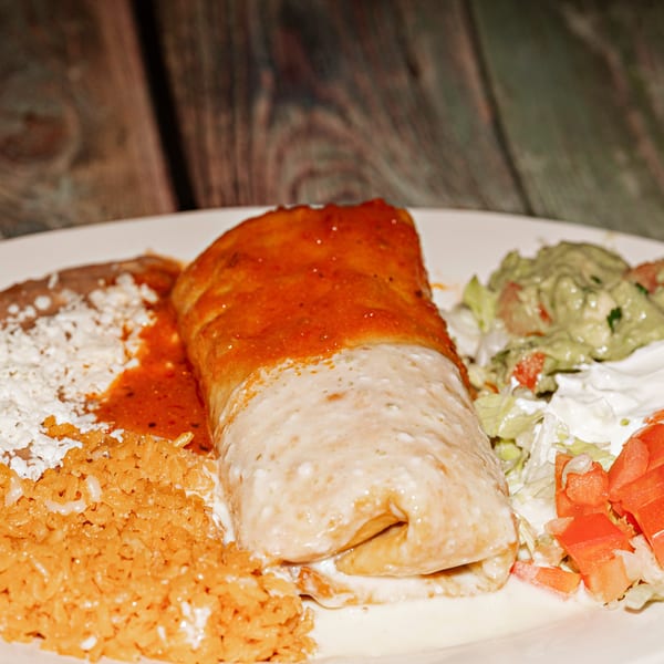 A burrito covered in red and white sauce, with rice, beans, guacamole, and salad on a white plate.
