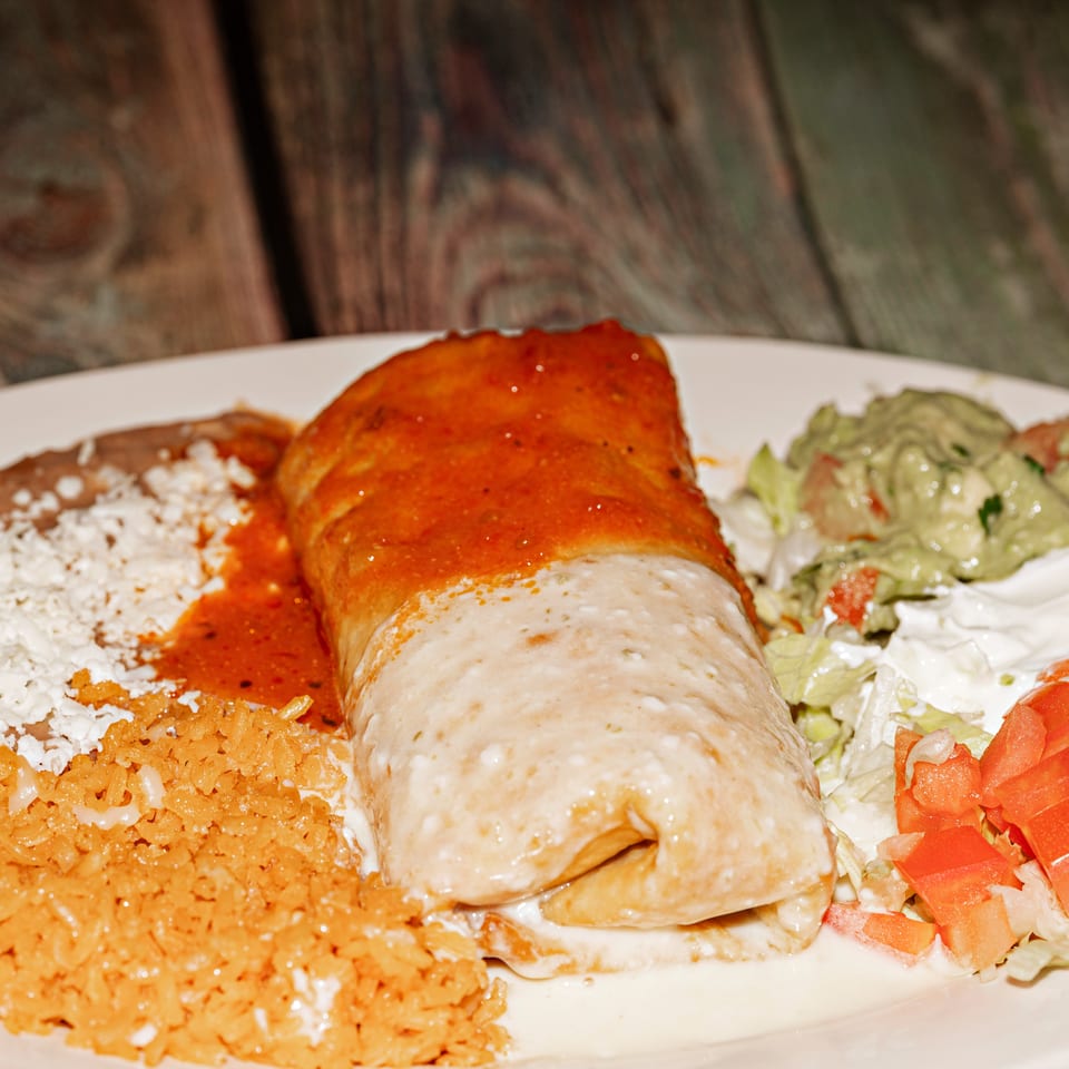 A burrito covered in red and white sauce, with rice, beans, guacamole, and salad on a white plate.