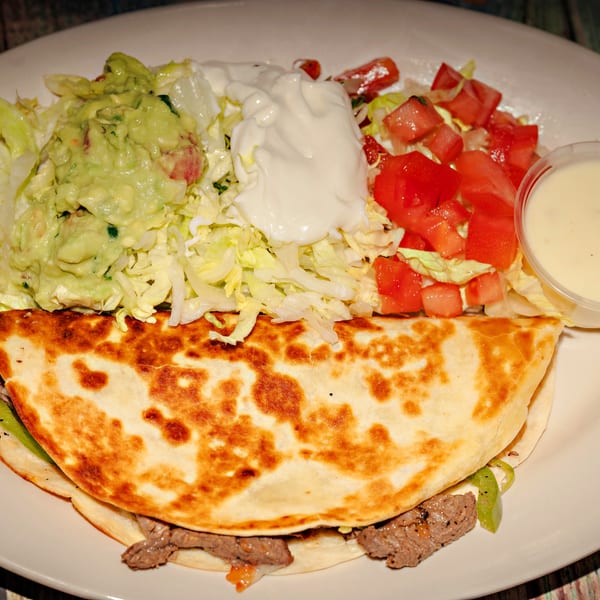 A beef quesadilla with guacamole, sour cream, diced tomatoes, shredded lettuce, and a side of sauce.